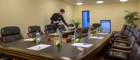 Un salón de conferencias con una larga mesa, sillas, botellas de agua, manzanas y una computadora portátil sobre la mesa.