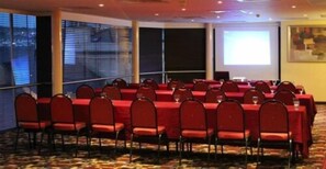 A conference room with rows of tables and chairs, large windows, and a projector screen.