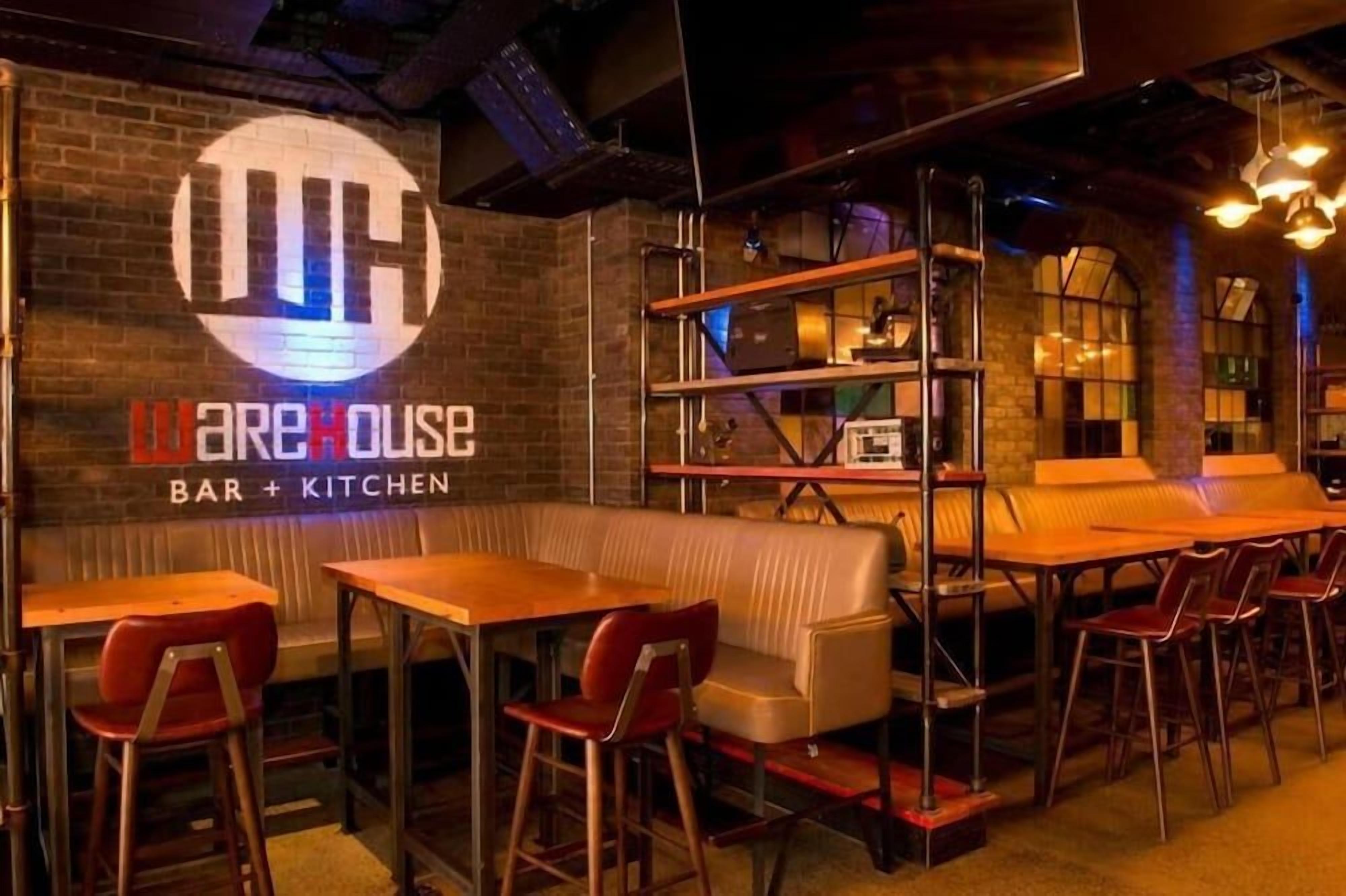 A restaurant interior with a bar and kitchen area, wooden tables, red chairs, and a brick wall with a logo.