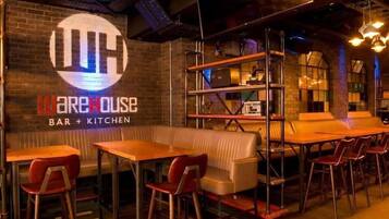 A restaurant interior with a bar and kitchen area, wooden tables, red chairs, and a brick wall with a logo.