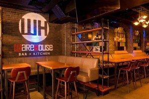A restaurant interior with a bar and kitchen area, wooden tables, red chairs, and a brick wall with a logo. - Mcgettigans Hotel Letterkenny (Letterkenny)