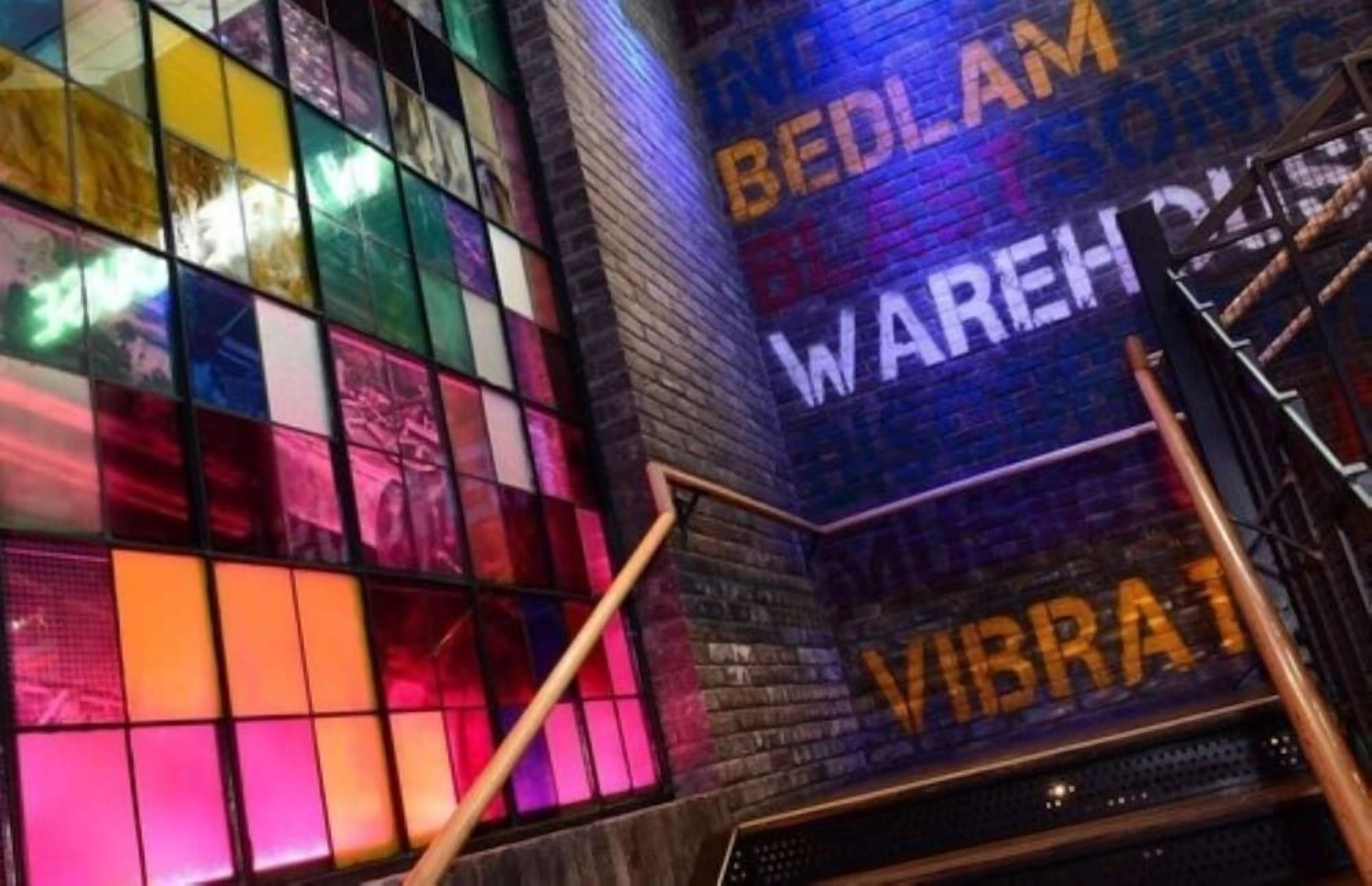 A modern interior with a colorful stained glass window and a staircase leading to a platform with a sign that reads 'BEDLAM WAREHOUSE.'