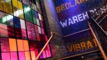 A modern interior with a colorful stained glass window and a staircase leading to a platform with a sign that reads 'BEDLAM WAREHOUSE.'