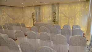 Una habitación con filas de sillas cubiertas de tela blanca, un escenario con cortinas y una iluminación decorativa.