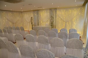 Una habitación con filas de sillas cubiertas de tela blanca, un escenario con cortinas y una iluminación decorativa.