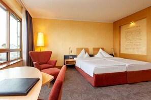 A hotel room with a bed, a desk, a chair, a lamp, and a window. - Vienna House by Wyndham Remarque Osnabruck (Osnabrueck)