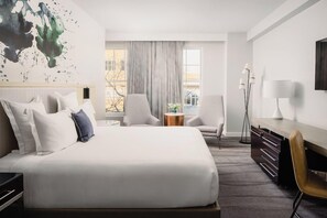 Habitación de hotel con una cama grande, un escritorio, una silla, una mesita redonda y un amplio ventanal con cortinas.
