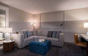 A modern living room with a grey sectional sofa, a blue ottoman, a standing floor lamp, and a framed artwork on the wall. - Kimpton Lorien Hotel and Spa (Alexandria)