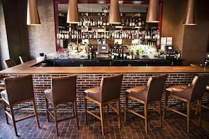 A bar area with a brick wall, wooden bar counter, leather bar stools, and a variety of bottles and glasses. - Kimpton Lorien Hotel and Spa (Alexandria)
