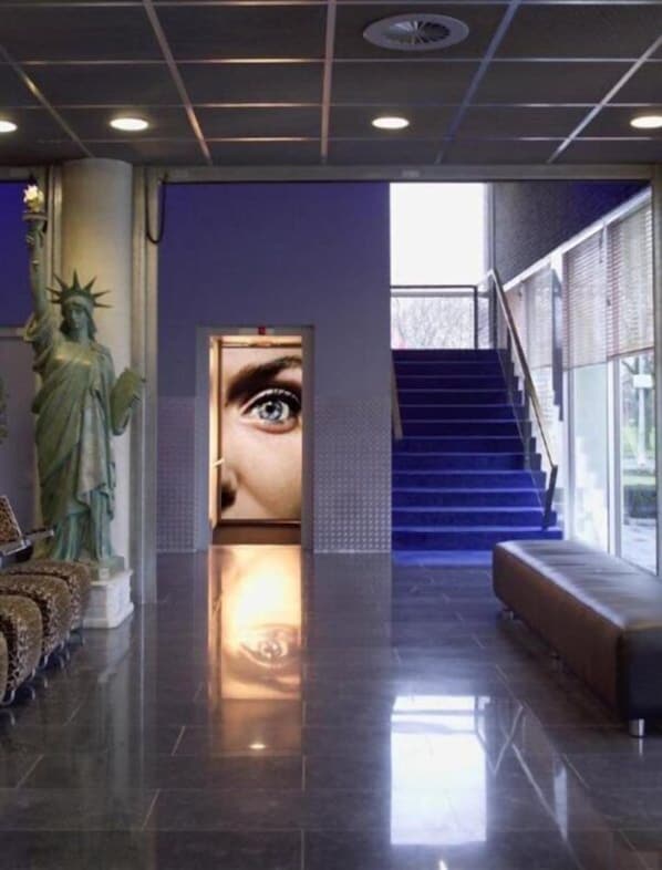 A lobby with a statue, a large digital display, a staircase, and a bench. - Select Hotel Apple Park Maastricht (Maastricht)