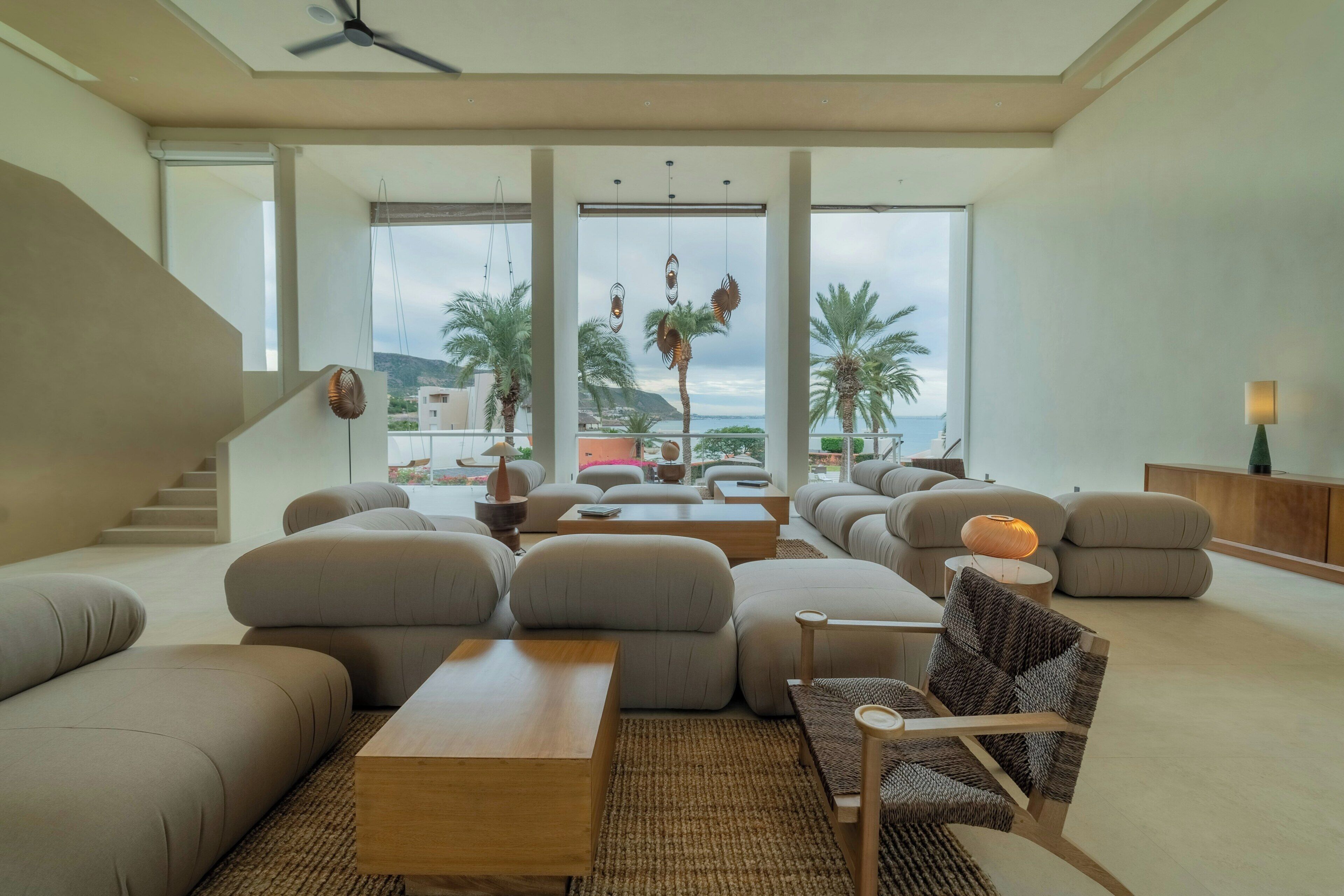 Un salón amplio con techos altos, ventanas grandes y vista a palmeras y un cuerpo de agua.