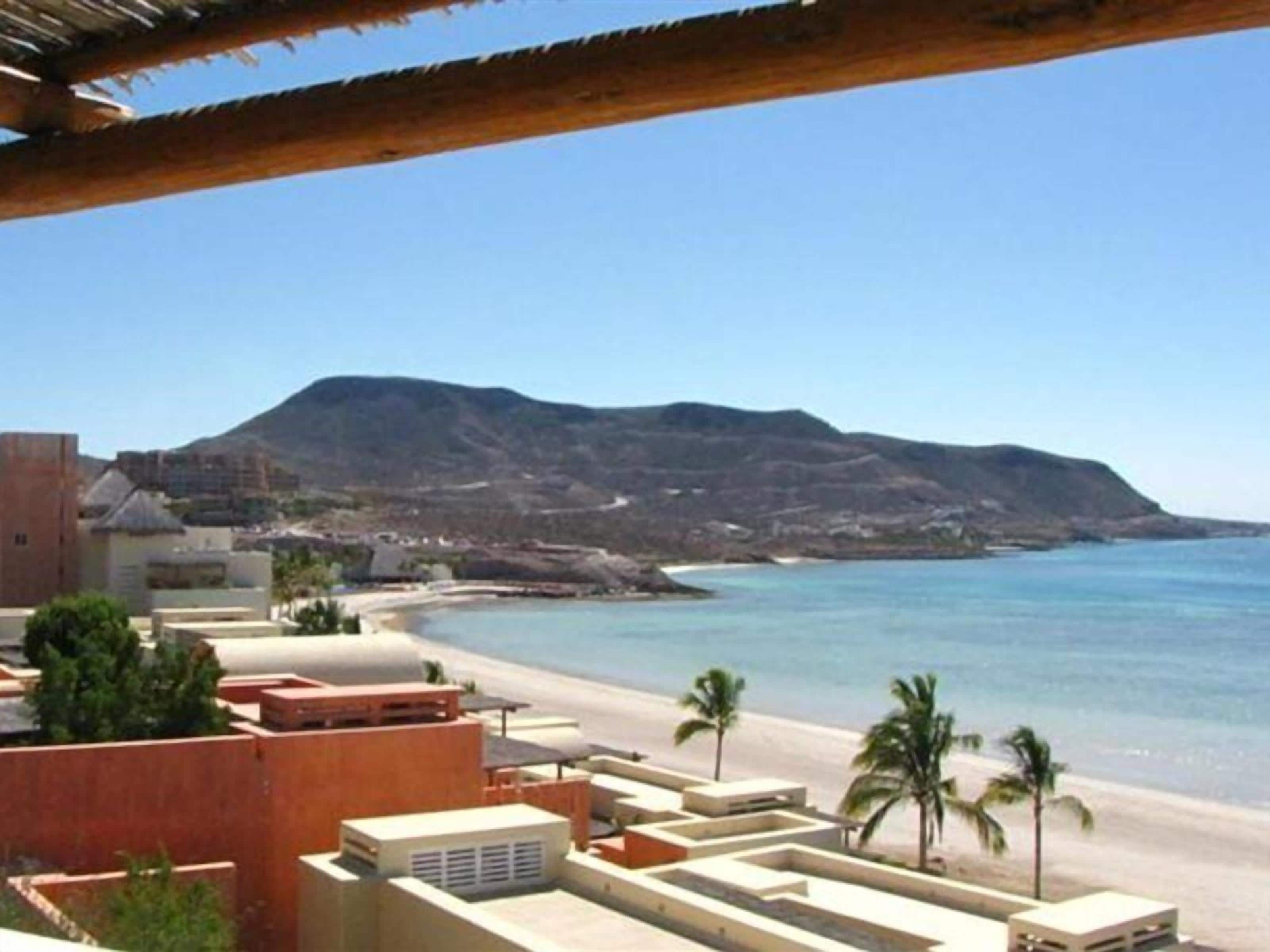 Um hotel à beira-mar com vista para a praia, uma montanha ao fundo e céu claro.