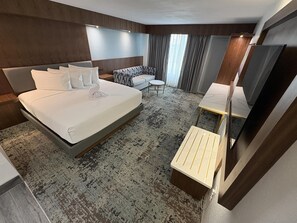 Quarto de hotel com uma cama grande, um sofá, uma mesinha, um espelho e uma televisão.