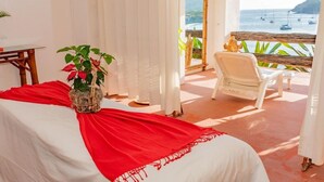 A room with a bed, a chair, a table, and a view of the sea.