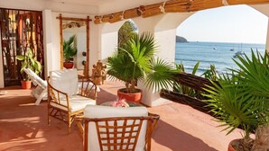 A covered outdoor seating area with wicker furniture, potted plants, and a view of the sea.