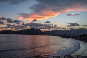 Paisaje costero con un atardecer, mar en calma y colinas a lo lejos.