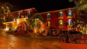A multi-story building with arched windows and balconies, surrounded by palm trees and illuminated by warm lighting.