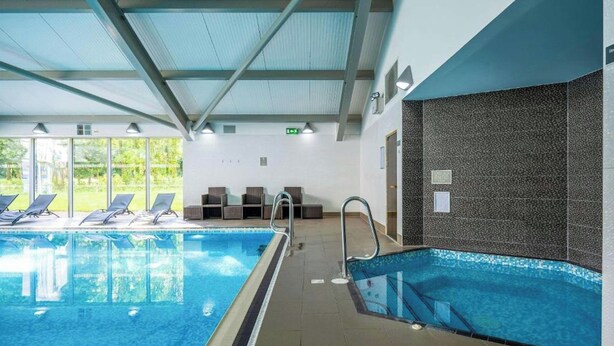 Una piscina interior con paredes de vidrio transparente, suelo de baldosas y techo con vigas de metal.