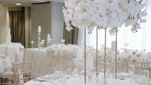 Un salón de banquetes con mesas redondas dispuestas para un evento formal, con arreglos florales blancos y centros de mesa con velas.