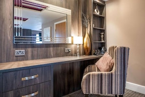 Ein Hotelzimmer mit einem hölzernen Waschtisch, einem Spiegel, einer Lampe, einem Stuhl und einem Regal mit Dekoration.