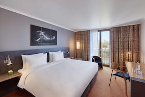 A hotel room with a large bed, a desk, a chair, a nightstand, a lamp, and a window with curtains. - Lindner Hotel Leverkusen Bayarena, Part of Jdv by Hyatt (Leverkusen)