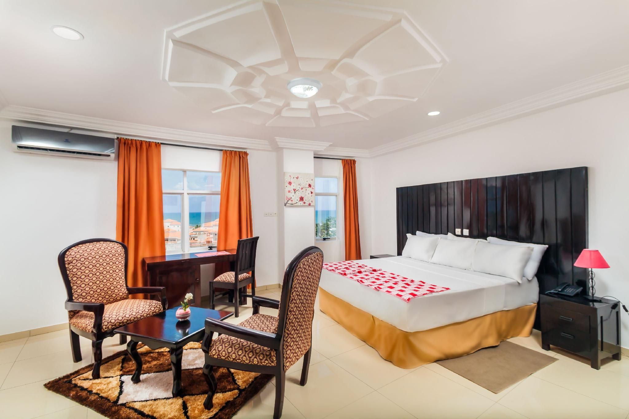 A hotel room with a bed, a desk, two chairs, a table, a nightstand, and a window with curtains.