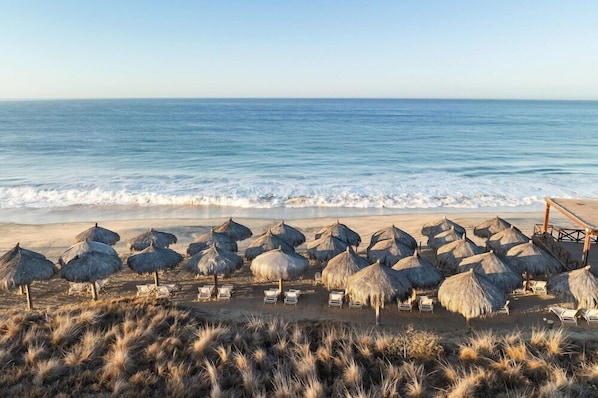 Una playa con sombrillas de paja y sillas de descanso, vista despejada del océano y un primer plano arenoso.