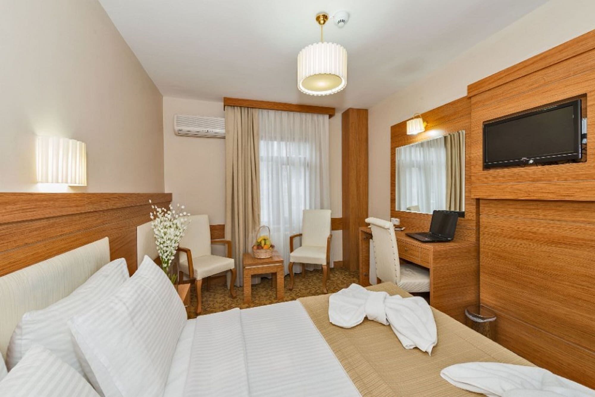 A hotel room with a bed, a desk with a laptop, a chair, a TV, and a window with curtains.
