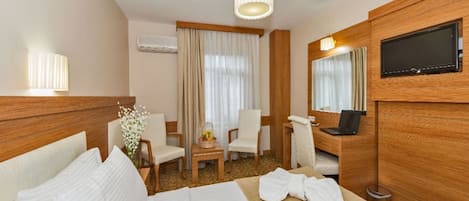 A hotel room with a bed, a desk with a laptop, a chair, a TV, and a window with curtains.
