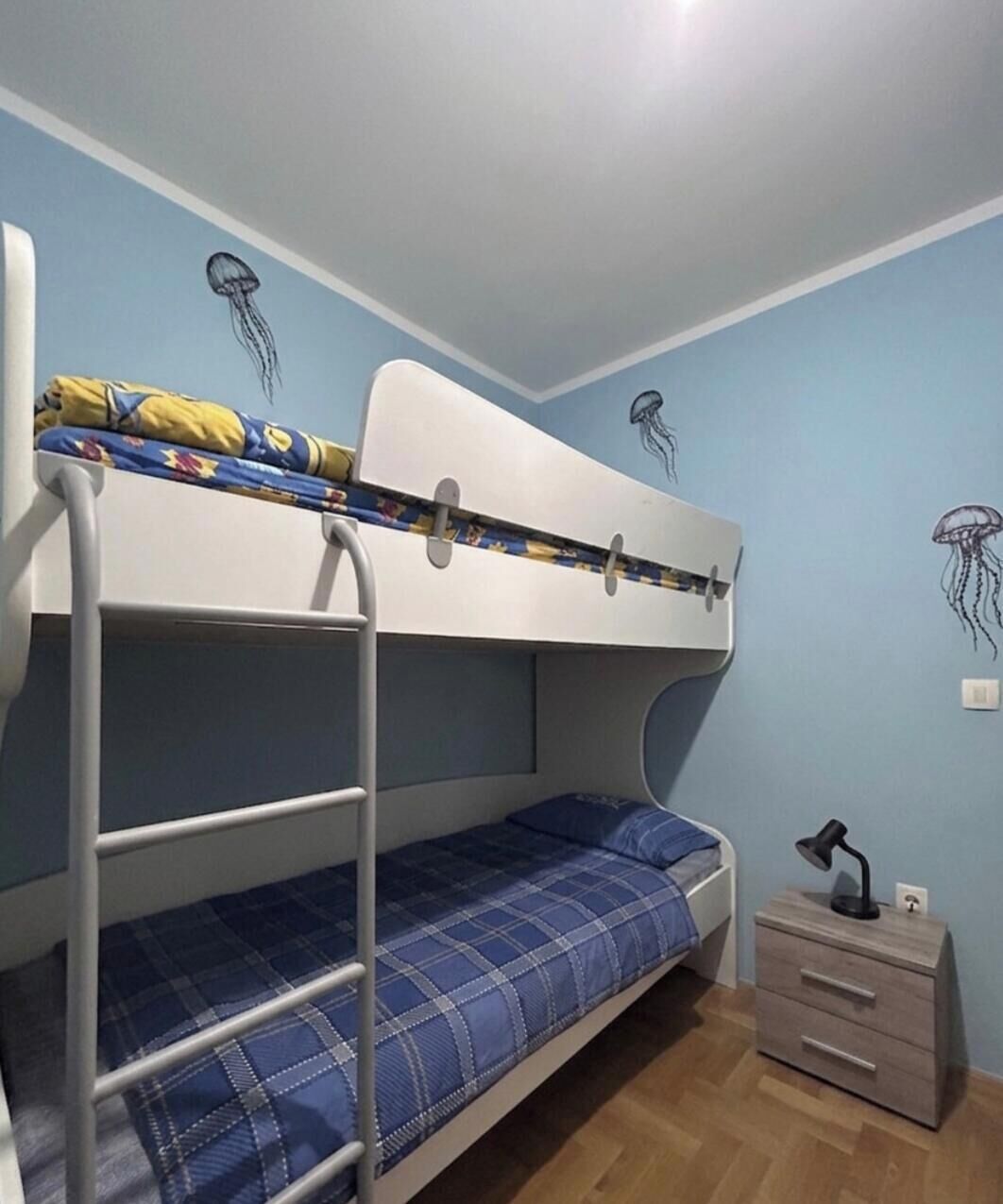 A bunk bed with a blue and yellow blanket, a small bedside table with a lamp, and a wall with jellyfish decals.