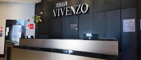 Una mesa de recepción con un cartel que dice 'GRAN VIVENZO' y una pizarra de anuncios con tarifas.