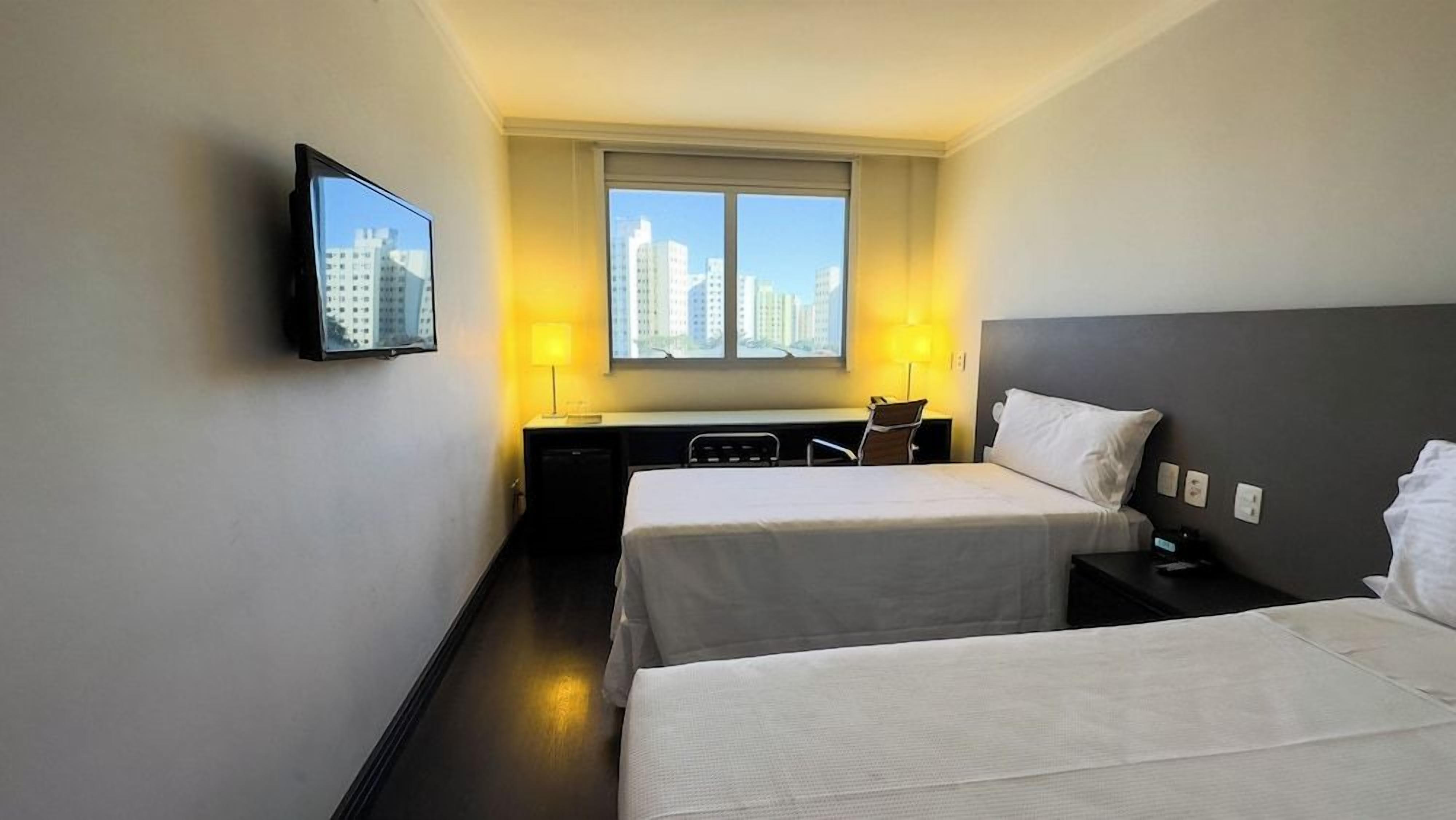 A hotel room with two beds, a desk, a chair, a TV mounted on the wall, and a window with a city view.