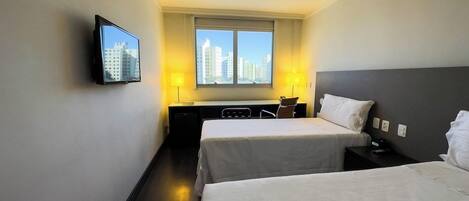 Quarto de hotel com duas camas, uma escrivaninha, uma cadeira, TV fixada na parede e janela com vista para a cidade.