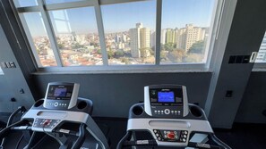 Two exercise bikes with digital displays, a window with a city view, and a control panel with buttons and a screen. - Hotel Gran Vivenzo Belo Horizonte (Belo Horizonte)