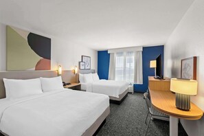 Habitación de hotel con dos camas, un escritorio, una silla, una lámpara y obras de arte en las paredes.