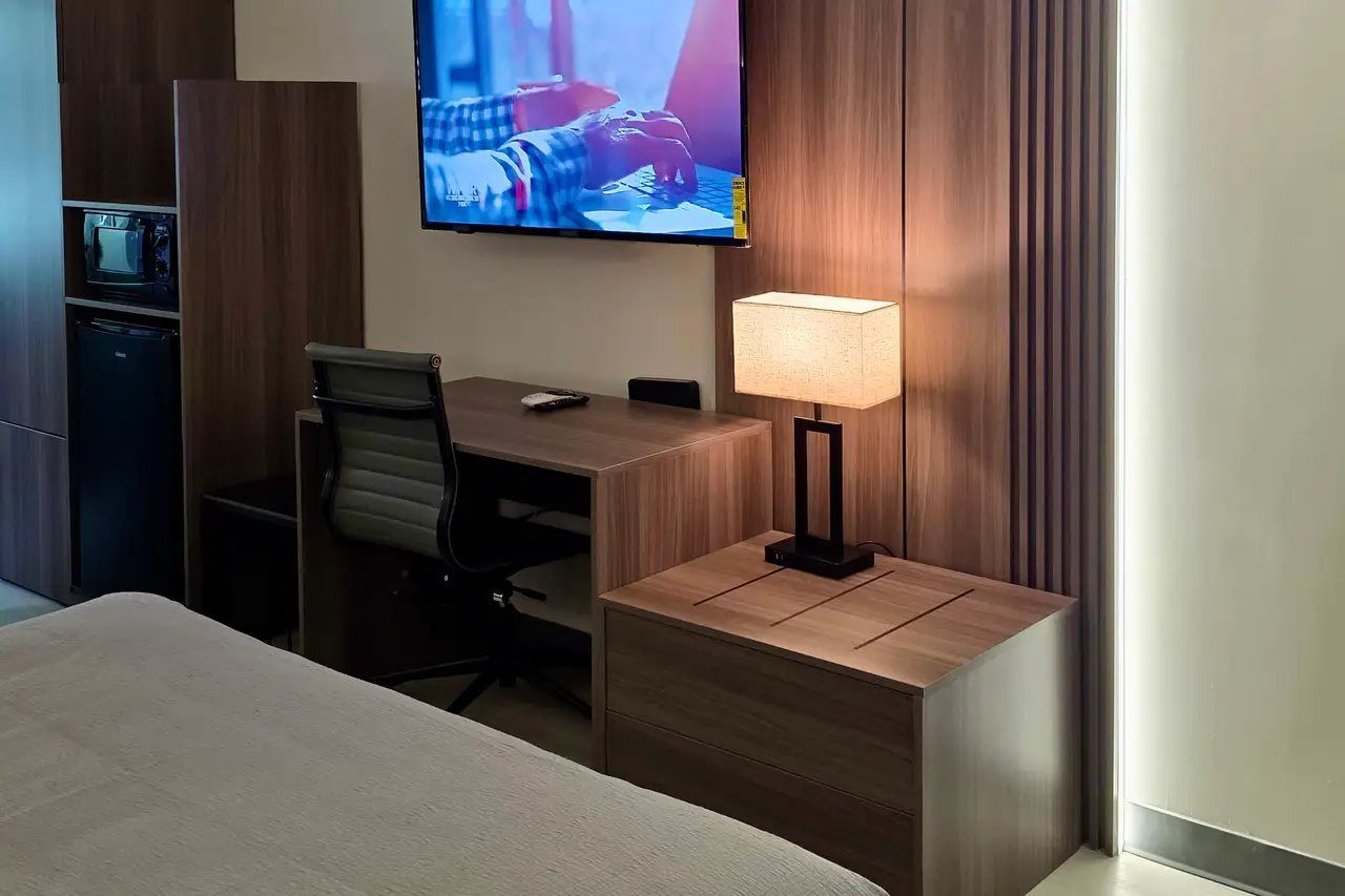 Ett hotellrum med en säng, ett skrivbord med en stol, en tv som är monterad på väggen och en sänglampa.