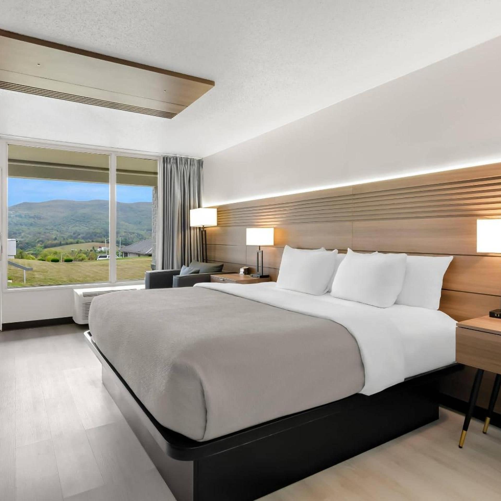 A modern hotel room with a large bed, bedside lamps, a view of the outdoors, and a wooden headboard.