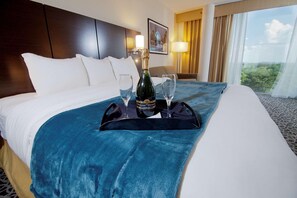 Habitación de hotel con cama, mesita de noche, lámpara, ventana con cortinas y vistas al exterior.