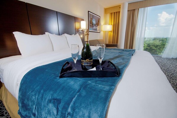 A hotel room with a bed, a nightstand, a lamp, a window with curtains, and a view of the outdoors. - Garden Plaza Hotel (Marietta)