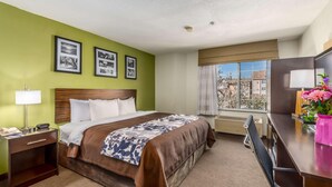 A hotel room with a large bed, a desk, a chair, a lamp, and a window with a view of buildings. - Rodeway Inn Airport Albuquerque, NM (Albuquerque)
