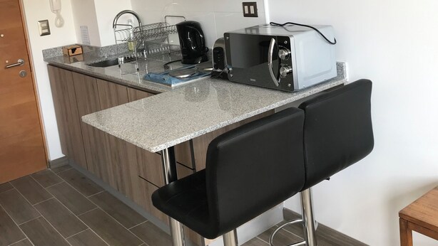 Cocina compacta con microondas integrado, tostadora y hervidor, un taburete pequeño y una puerta de madera.