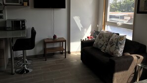 Una sala de estar moderna con un televisor de pantalla plana, un sofá con cojines estampados, una zona de comedor con microondas y una ventana con vistas a edificios.