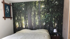 Un dormitorio con un gran tapiz de temática forestal, una cama con cubrecama blanco texturizado, una mesita de noche con lámpara y un pequeño cuadro en la pared.