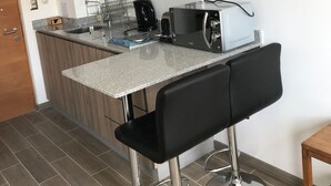 Cocina compacta con microondas, tostadora y cafetera sobre la encimera, un mueble de madera y una silla negra con base metálica.