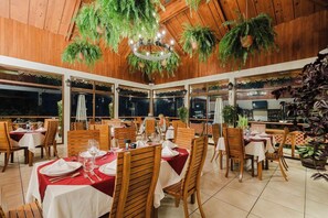An indoor dining area with wooden tables and chairs, large windows, and hanging plants. - Hotel Ficus Monteverde (Monteverde)