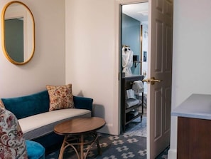 A hotel room with a sofa, a round wooden table, a mirror, and a door leading to a bathroom with a towel rack and a robe. - Artmore Hotel (Atlanta)