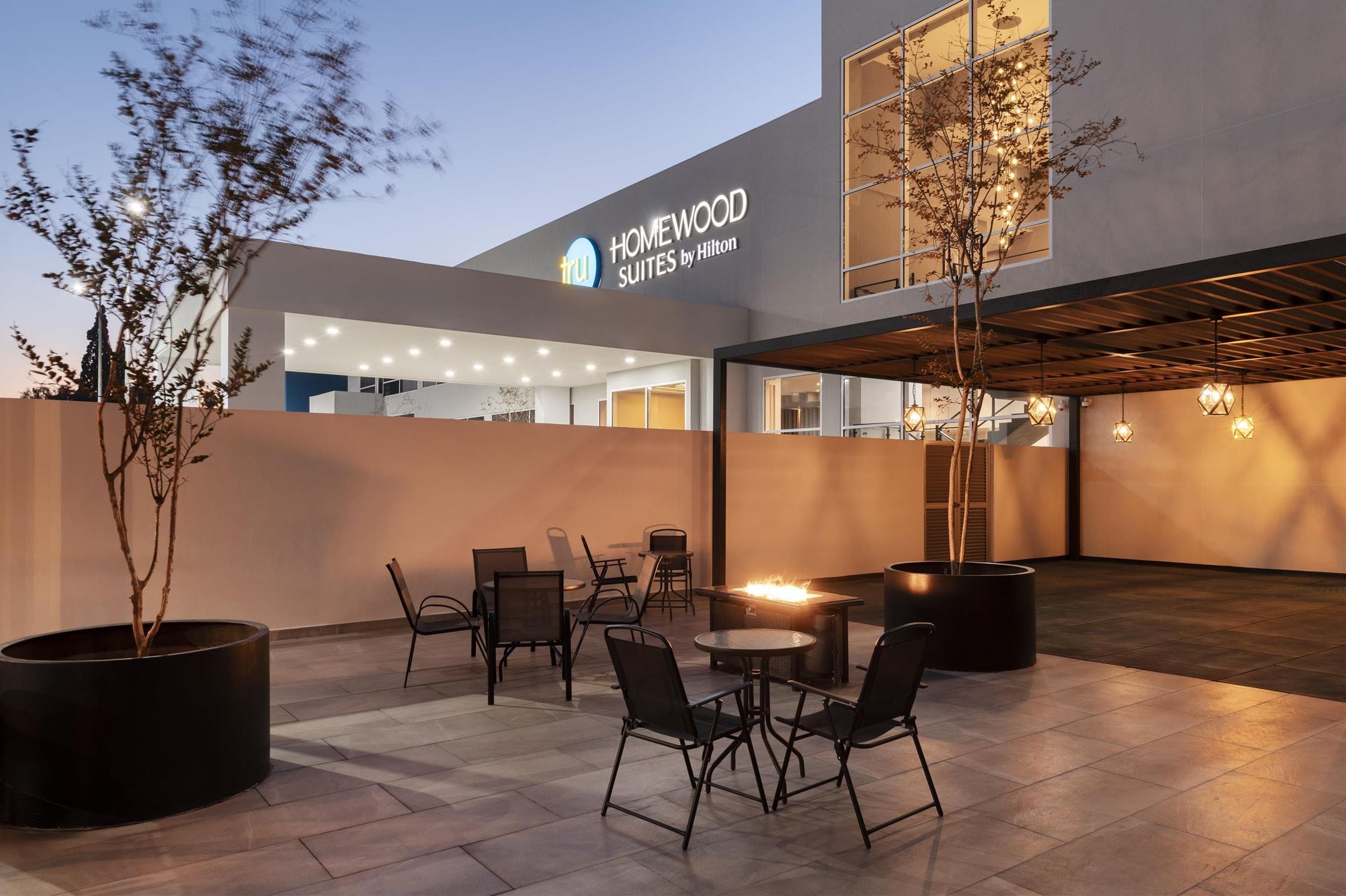 Exterior de un hotel con zona de estar al aire libre, chimenea y un letrero que dice 'HOMEWOOD SUITES by Hilton'.