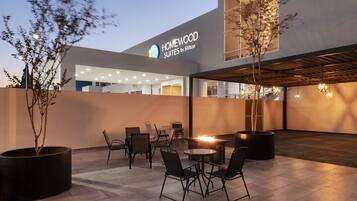 Exterior de un hotel con zona de estar al aire libre, chimenea y un letrero que dice 'HOMEWOOD SUITES by Hilton'.