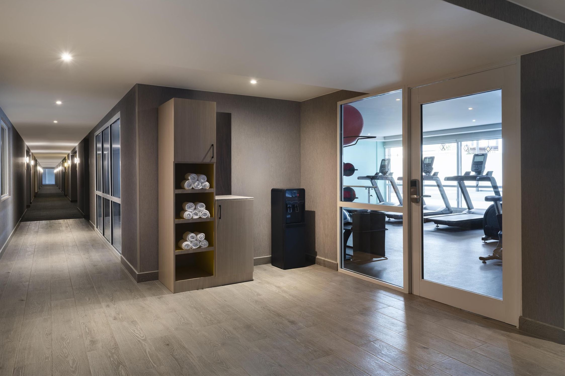 Un pasillo moderno de hotel con un gimnasio visible a través de una puerta de vidrio.