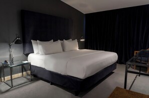 Een moderne hotelkamer met een groot bed, een glazen bijzettafeltje, een bureau en een donker gordijn.